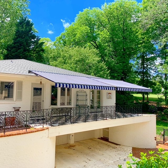 Wide blue and white striped retractable awning spanning the full width of a white brick home with ornamental iron railing patio and lush green trees